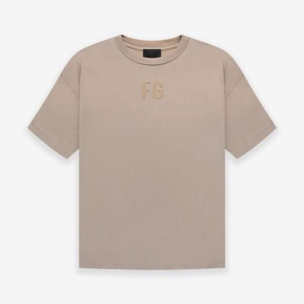 A Fear of God Essentials FG T-shirt in 'Dusty-Beige' featuring short sleeves and a minimalist design. The letters FG are subtly printed on the chest in a slightly darker hue. The t-shirt is displayed flat against a plain white background.