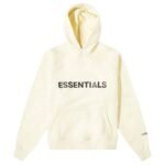 The Fear of God Essential Hoodie in 'Buttercream' is cream-colored with bold black ESSENTIALS lettering on the front. It includes a front pocket and hood, with smaller ESSENTIALS text printed on the left sleeve near the wrist.