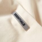 Close-up image of the Fear of God Essential Hoodie 'Buttercream' showcasing a beige fabric with a rectangular, raised label. The light gray label features the word ESSENTIALS in bold, black letters. The fabric's texture appears soft and smooth.