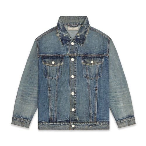 The Essentials Fear of God Denim Jacket is a blue design with a button-up front, featuring two chest pockets with buttoned flaps. It has a slightly faded and worn appearance, complemented by a pointed collar and long sleeves.