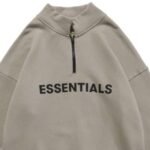 A beige Essentials Half Zip High Collar Loose Hoodie with "ESSENTIALS" printed in bold black letters across the chest, featuring a high collar and a simple, casual design.