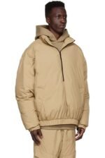 A person wearing the Essentials Tan Polyester Jacket, complete with a hood, stands against a white background. The jacket is paired with matching beige pants, creating an outfit that looks warm and suitable for cold weather.