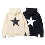 Two Fear Of God Essentials Star Hoodies are laid flat, one in beige with a black star on the back and the other in black with a white star. Both feature hoods and long sleeves, displayed side by side.