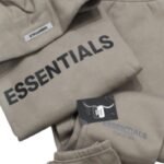 The Fear Of God Essential Hooded Tracksuit 'Brown' is on display, featuring beige clothing items like a sweatshirt and sweatpants with the word ESSENTIALS emblazoned in bold black letters and a small logo tag visible on the fabric.