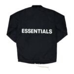 The Fear of God Essentials Coach Jacket is black with the word "ESSENTIALS" prominently printed in large white letters on the back. It features a simple design complemented by drawstrings at the bottom.